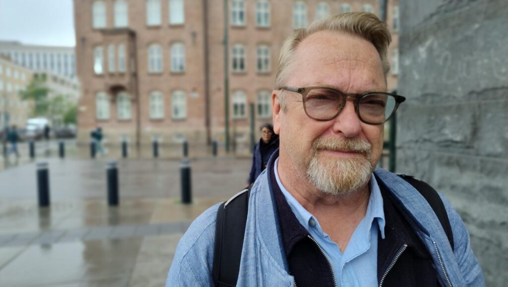 Porträtt på en man i 60-årsåldern med glasögon och skägg framför en tegelbyggnad i stadsmiljö.