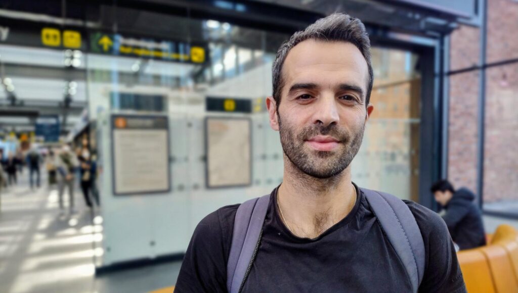 Porträtt på en man i 35-årsåldern med ryggsäck i en ljus stationsmiljö med informationsskyltar i bakgrunden