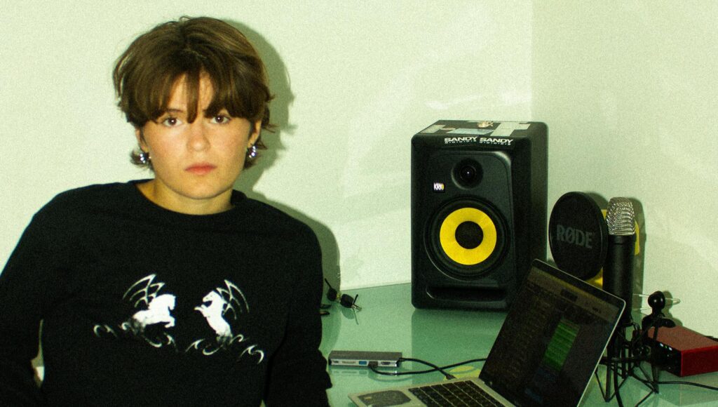 Kvinna i 20-årsåldern med kort brunt hår sitter vid ett skrivbord med laptop, studiomonitor och mikrofon.