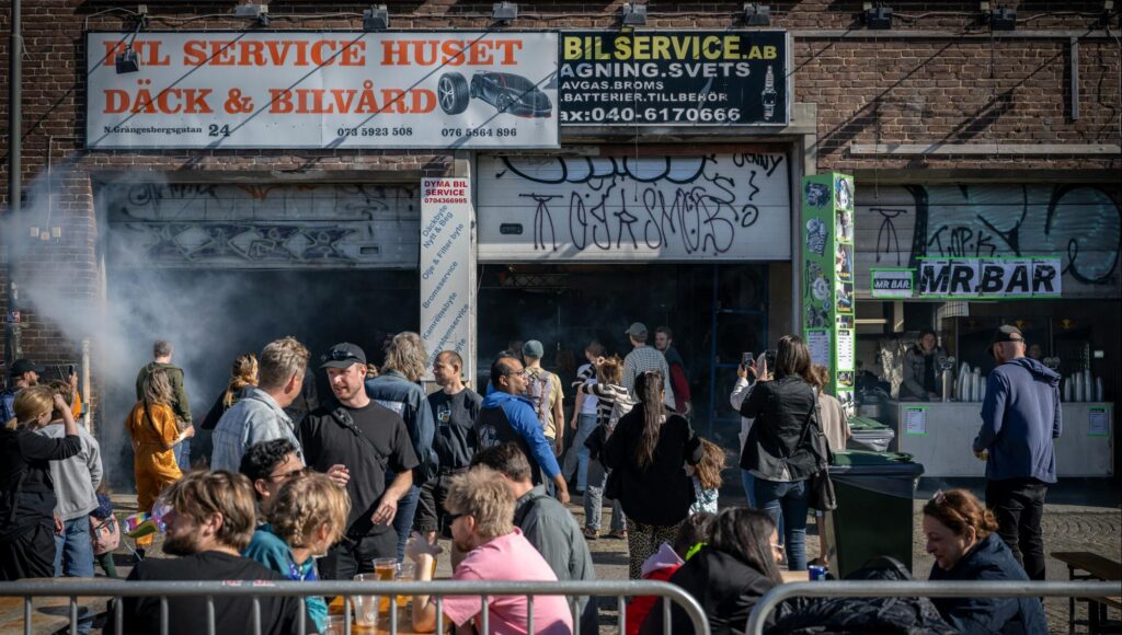 Folkmassa samlad vid en graffitimålad verkstadslokal med rök och skyltar under en gatufest.