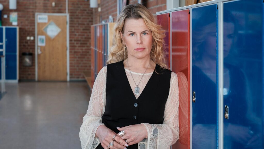 Blond kvinna i svart väst och spetströja står framför röda och blå elevskåp i en skolkorridor med tegelvägg.