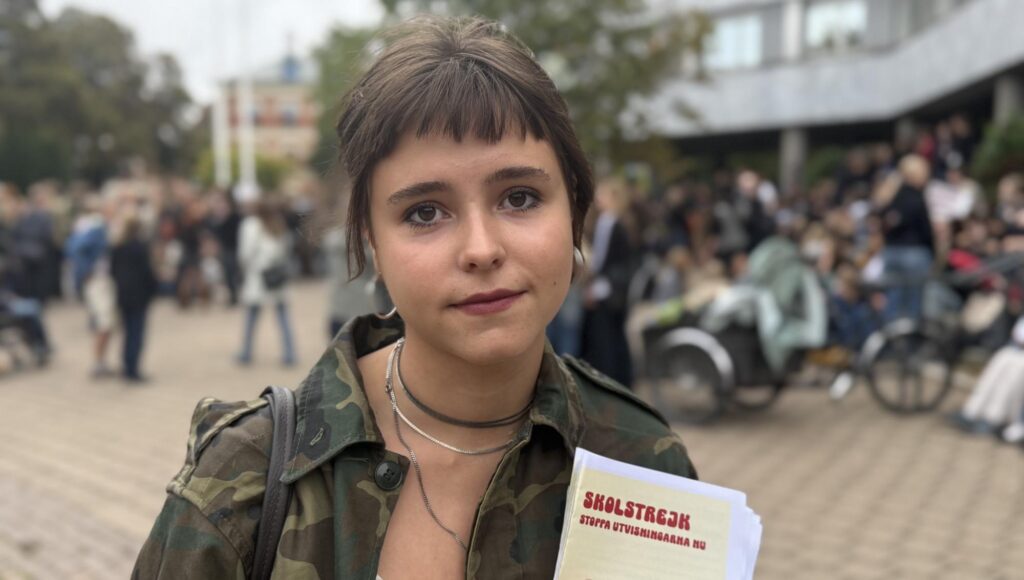 Ung kvinna i kamouflagejacka håller i ett flygblad med texten Skolstrejk framför en folksamling utomhus.