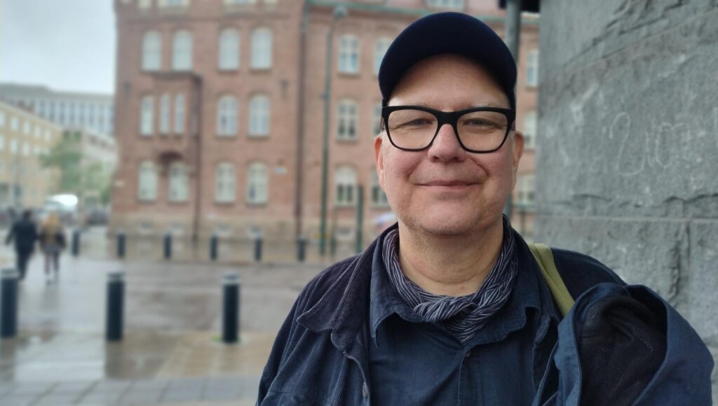 En man i glasögon och keps ler mot kameran utomhus i stadsmiljö med en tegelbyggnad i bakgrunden.