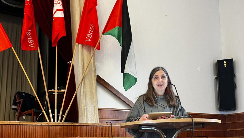 En kvinna i 50-årsåldern talar vid ett podium bredvid Vänsterpartiets flaggor och den palestinska flaggan.