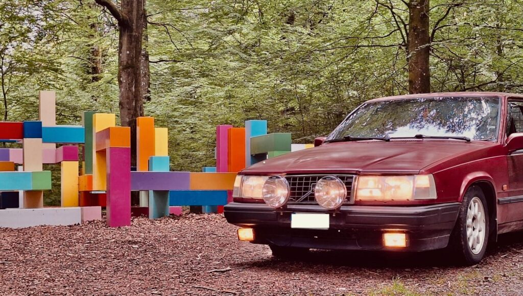 Röd Volvo 940 med extraljus framför en abstrakt, färgglad träskulptur i en grön lövskog.