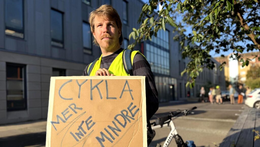 Man i reflexväst håller en skylt med texten Cykla mer inte mindre framför en modern byggnad.
