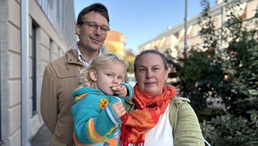En man, en kvinna och ett litet barn står tätt tillsammans på en gata i solljus.