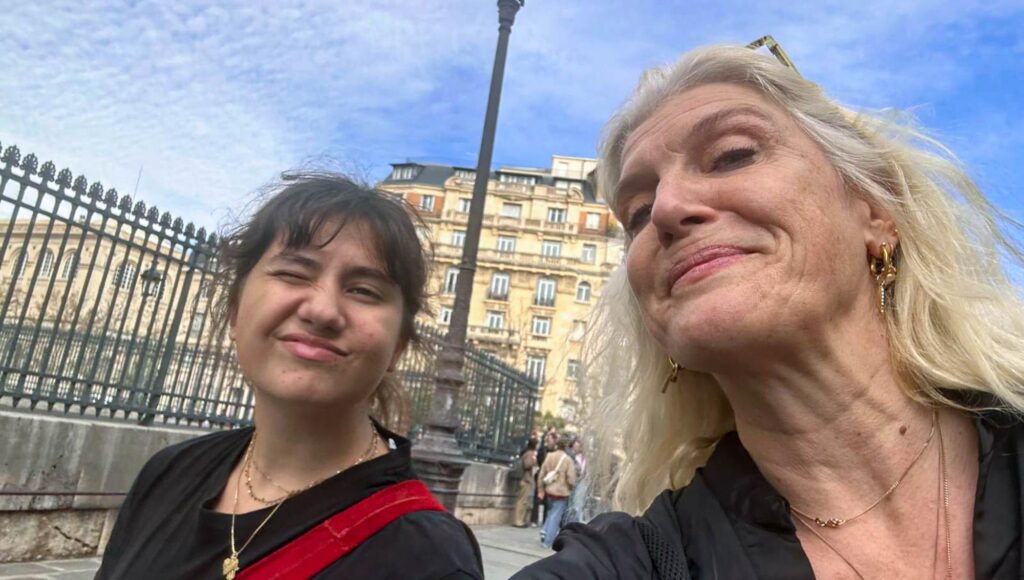 Närbild på en leende kvinna med grått hår och en blinkande ung kvinna framför klassiska byggnader i Paris.