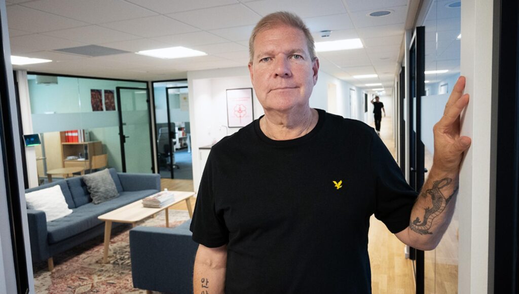 Man i svart t-shirt med tatueringar står i en kontorskorridor med soffgrupp i bakgrunden.