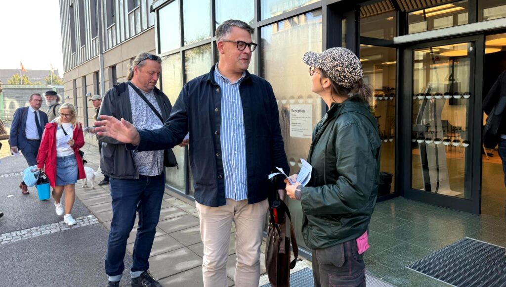 En man i glasögon och blå jacka samtalar med en kvinna i keps framför en entré med glasdörrar.