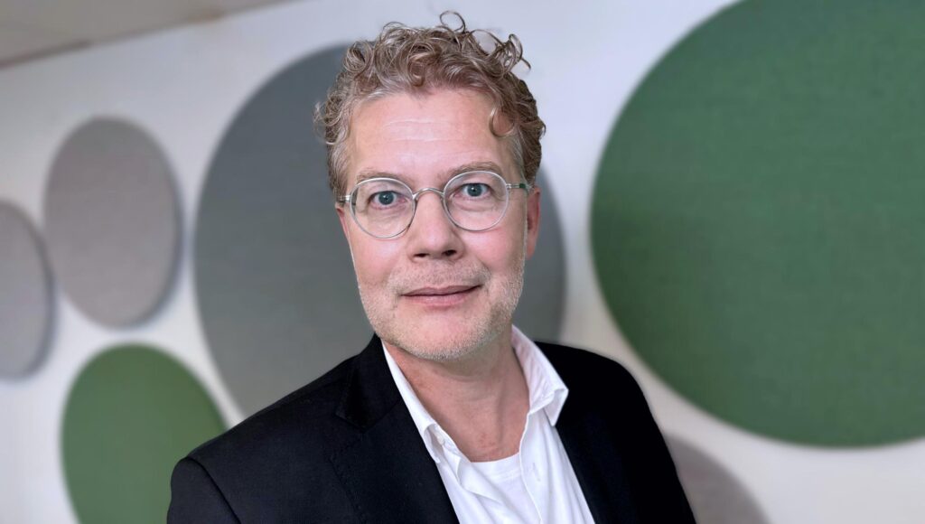 Porträtt av en man med lockigt hår och runda glasögon framför en vägg med gröna och grå cirklar.