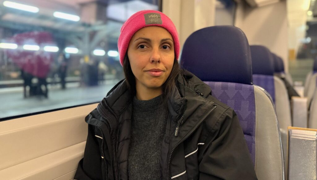 Kvinna i 30-årsåldern med rosa mössa och svart jacka sitter på ett tåg.
