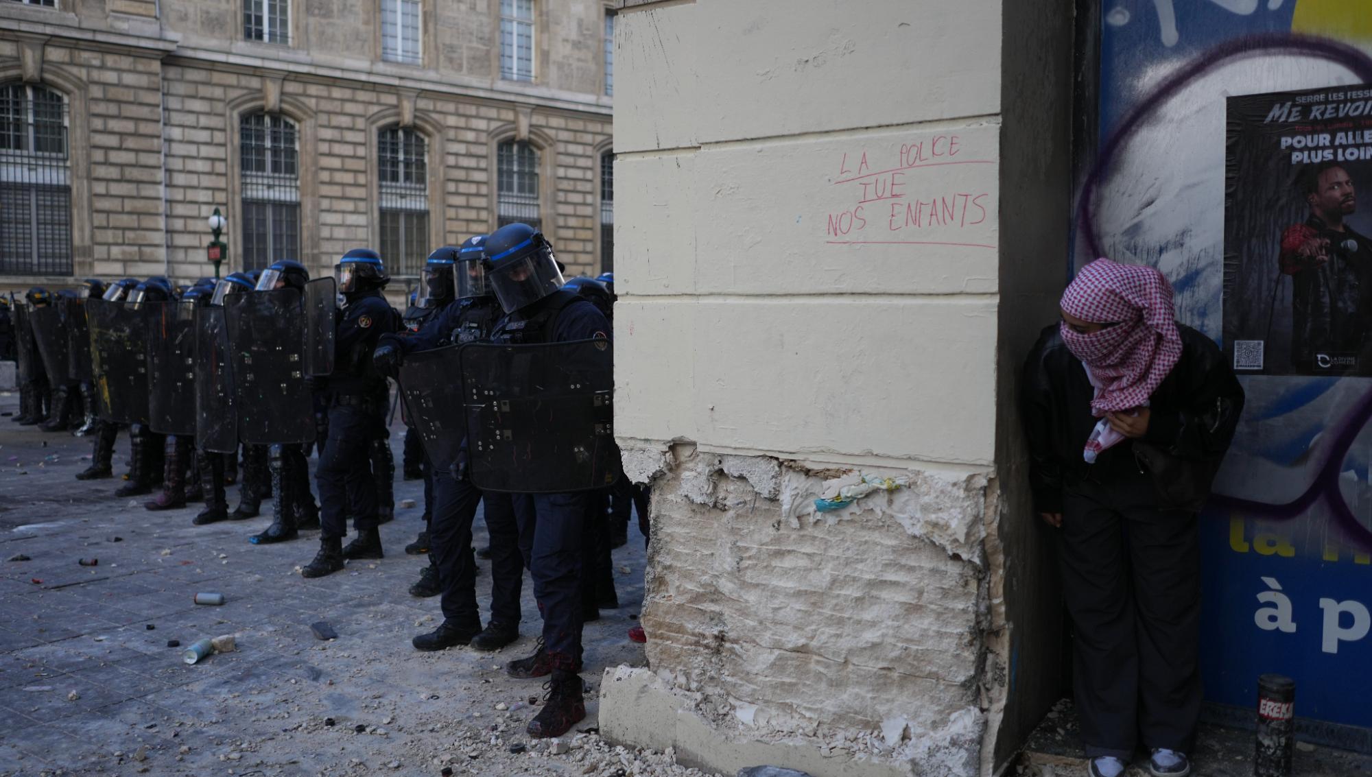 En demonstrant betraktade kravallpoliser intill en vägg med budskapet ”Polisen dödar våra barn” under helgens våldsamma protest i Paris. Demonstrationen hölls mot stigande levnadskostnader och till stöd för studenter som protesterat mot bristande resurser i skolan. Foto: Thibault Camus /AP/TT