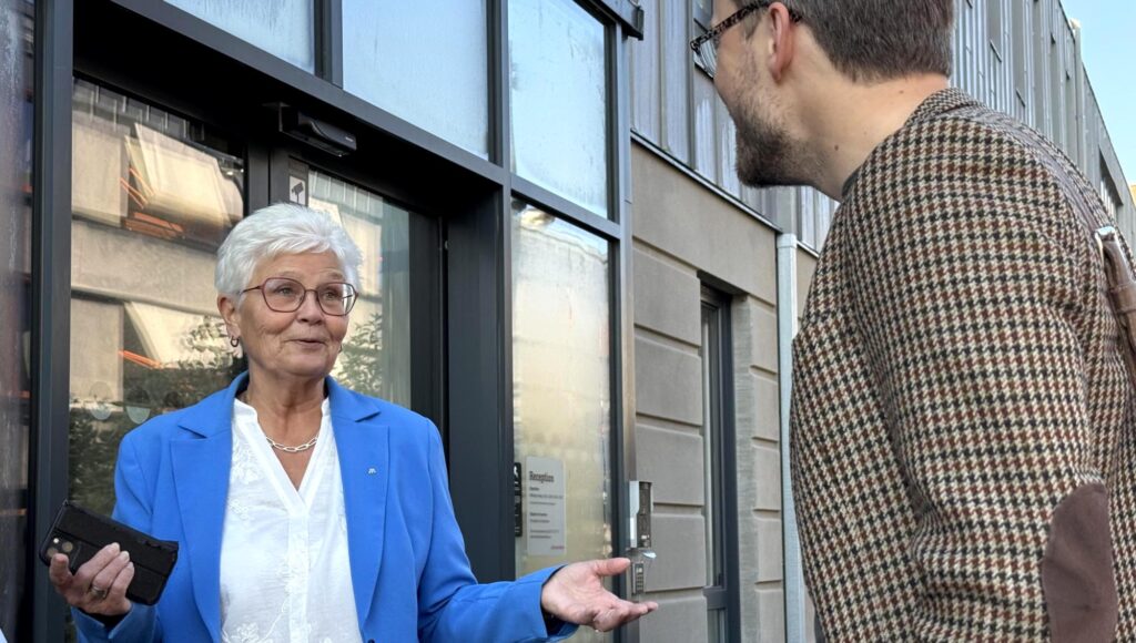 Kvinna i blå kavaj och man i mönstrad kavaj samtalar framför en glasdörr till en modern byggnad.