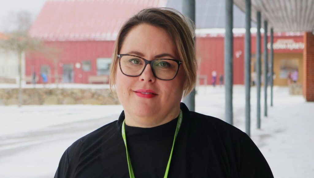 Kvinna i glasögon ler mot kameran utomhus framför en röd skolbyggnad i vintrigt landskap.